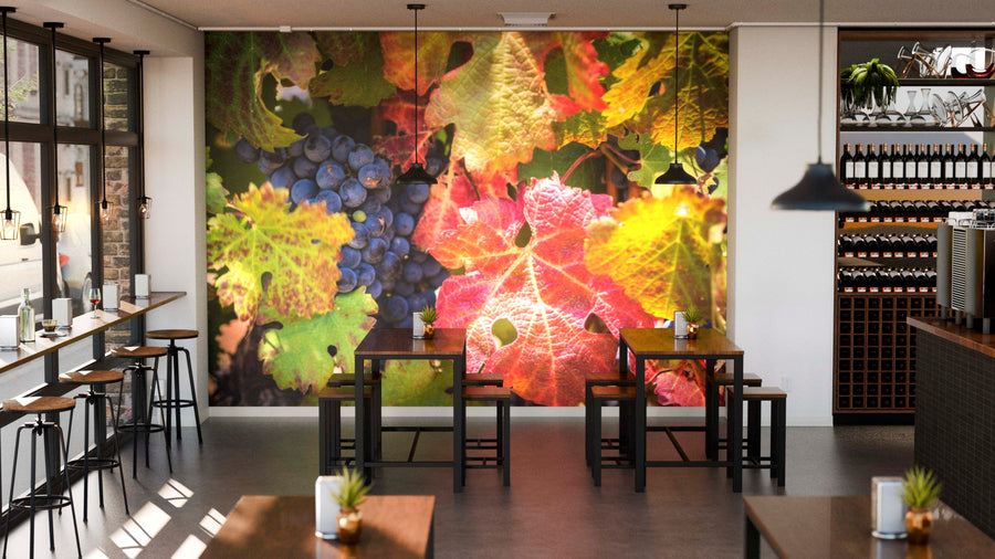 A cozy modern café with wooden tables, potted plants, and large windows. The back wall features the Vineyard Wall Mural of grape clusters and autumn leaves, adding warmth. Wine bottles are displayed on shelves.