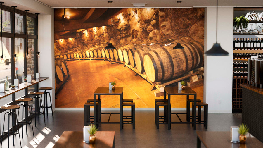 Modern wine bar with high stools, wooden tables, and potted plants. The back wall features the Vintage Wine Barrels Wall Mural depicting a cozy cellar scene. Glass windows line the left; shelves of wine bottles are visible on the right. Warm, inviting vibe.