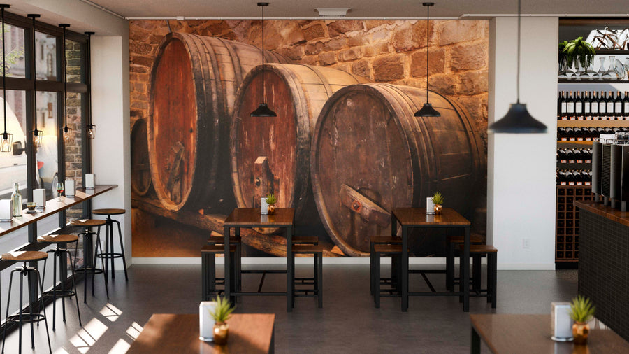 A cozy cafe with wooden tables, stools, and potted plants features the Vintage Wooden Barrels Wall Mural on a stone wall. Pendant lights hang above, and large windows fill the space with natural light.