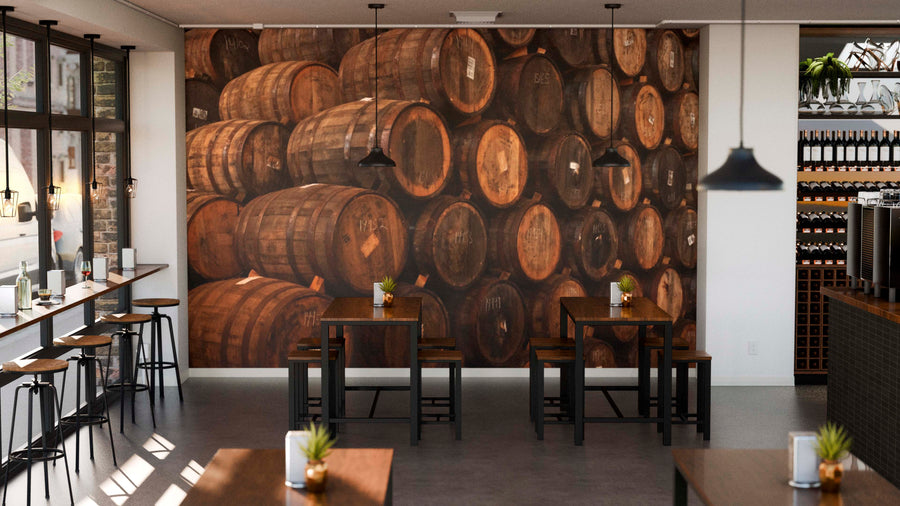 A cozy café interior with wooden tables and chairs, potted plants, and a Whiskey Barrel Storage Wall Mural as a feature wall. Pendant lights hang overhead, while sunlight pours in from large windows on the left side.