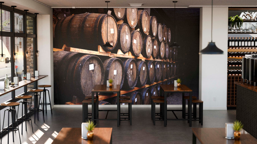 A modern café with wooden tables, black chairs, potted plants, and a Wine Barrel Collection Wall Mural. Soft ceiling lights illuminate shelves with bottles on the right, while large windows fill the space with natural light.