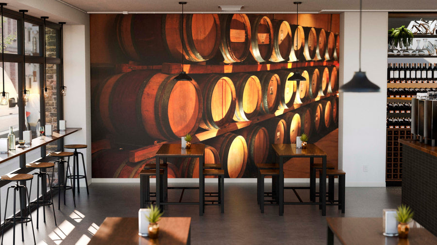 A cozy cafe features wooden tables, stools, small plants, and a warm atmosphere with sunlight from large windows. The highlight is the Wine Barrels Wall Mural depicting rows of wine barrels on the main wall.