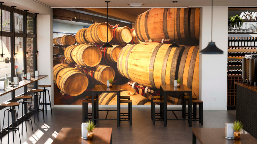 A cozy modern cafe with wooden tables, barstools by a window, and a large Wine Barrels Cellar Wall Mural. Pendant lights hang from the ceiling, potted plants decorate tables, and wine bottles are displayed on shelves.