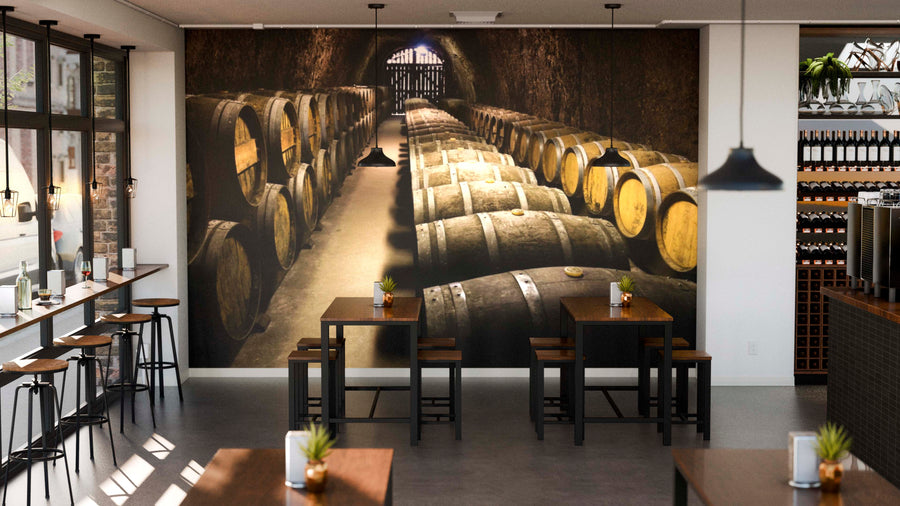 A modern café with wooden tables and potted plants has a custom "Wine Barrels in Cellar Wall Mural" on the back wall, bringing an inviting, rustic vibe with its depiction of rows of barrels.