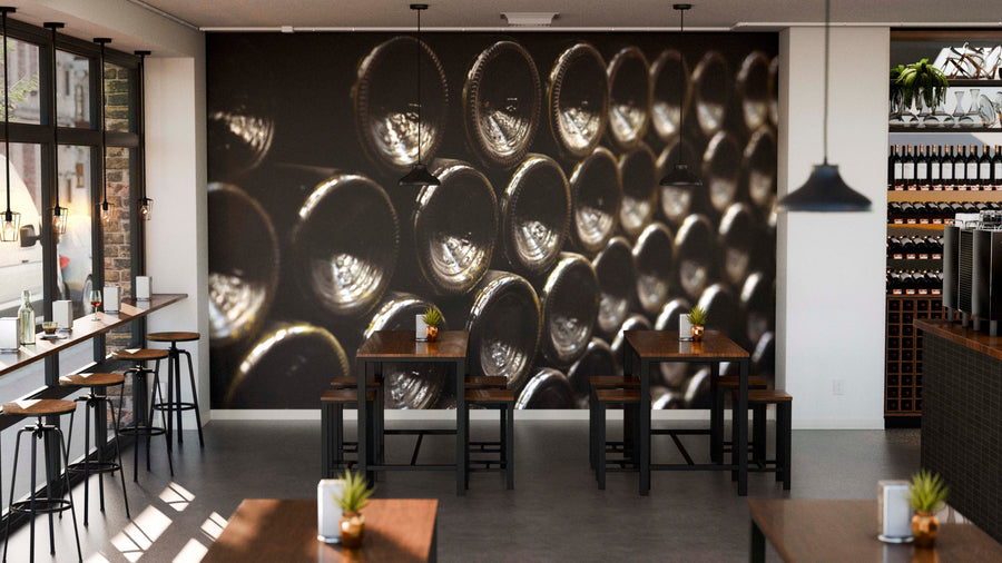 Modern cafe with wooden tables and black chairs. The back wall displays the Wine Bottles Resting Horizontally Wall Mural, showing stacked wine bottles from below. Natural light enters left; shelves with bottles line the right wall.