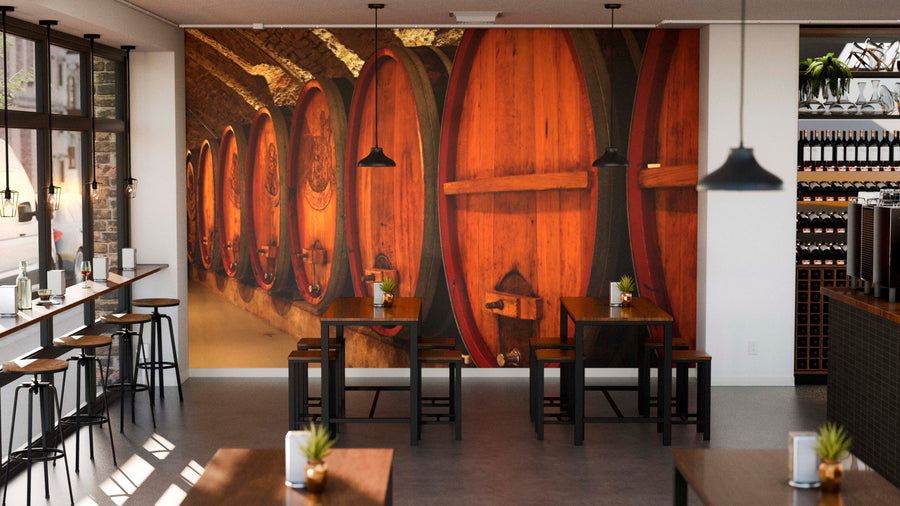 Modern café interior with wooden tables and black chairs. The back wall features the Wine Cellar Wall Mural, depicting rows of wine barrels. Pendant lights hang above, windows line the left, and small plants accent each table.