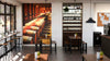 A modern wine bar with wooden tables, high stools by large windows, a tall wine rack, potted plants, pendant lights, a bulletin board, and a bright atmosphere features the Wine Cellar Interior Wall Mural depicting wine barrels.