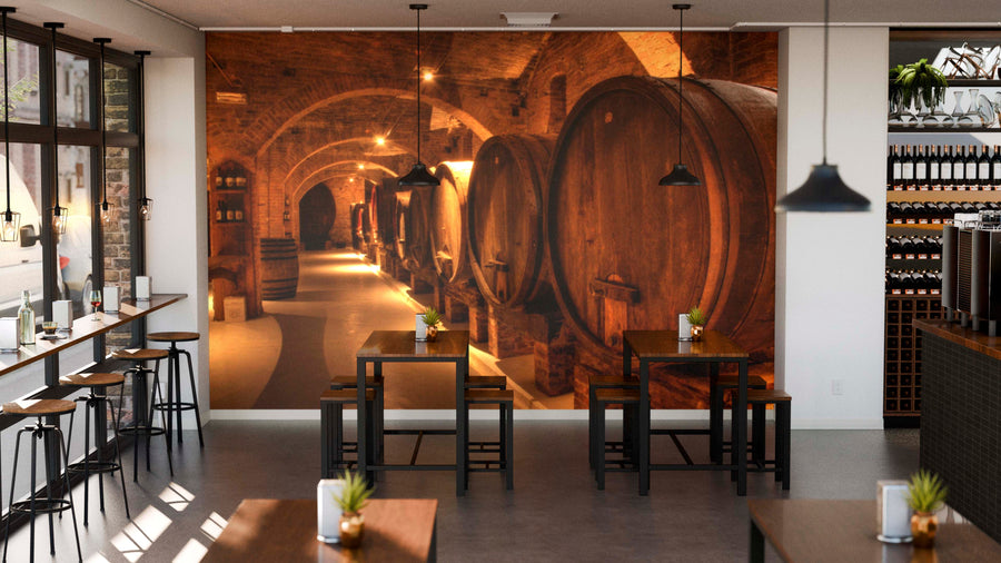 A modern café with high stools, wooden tables, and pendant lights. The back wall displays the Wine Cellar in Abbey Wall Mural, showing arched ceilings and large barrels. Small potted plants decorate each table by the windows.