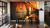 A stylish cafe with wooden tables and black chairs features the Winery Barrels Wall Mural. Pendant lights hang above, potted plants decorate tables, wine bottles line shelves, and large windows welcome natural light.