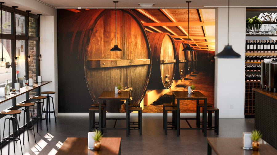 A stylish cafe with wooden tables and black chairs features the Winery Barrels Wall Mural. Pendant lights hang above, potted plants decorate tables, wine bottles line shelves, and large windows welcome natural light.