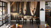 A cozy, modern café with wooden tables and stools, potted plants, sunlit windows, shelves of wine bottles and glasses, and a Wooden Cask Wall Mural as a peel-and-stick backdrop creating a warm, inviting atmosphere.