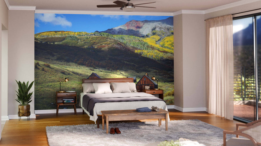 A bright bedroom features a bed with a wooden headboard, nightstands, a bench, and a Barns in San Juan Mountains Wall Mural. The room includes a rug, plant, ceiling fan, and glass doors with beige curtains streaming sunlight.
