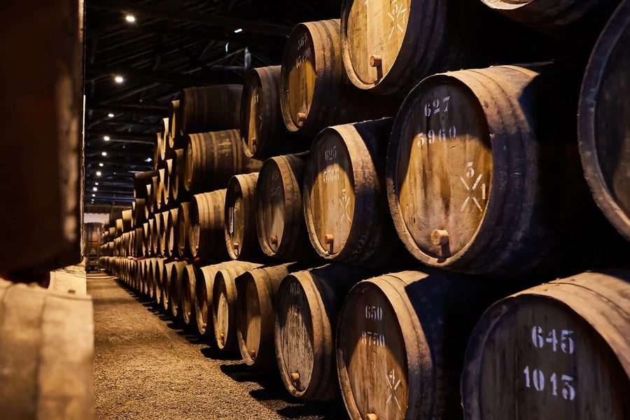 Modern café interior with wooden tables, black chairs, and large windows. The back wall displays the Barrel Storage Wall Mural showing stacked wine barrels. Shelves lined with wine bottles run along the right wall beneath pendant lights.