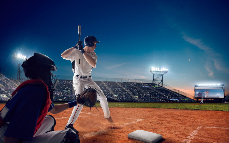 A bedroom features the Batter On-Deck Wall Mural with a baseball batter and catcher at a stadium. The room includes a bed, desk with computer, chair, wall art, sunlight through blinds, and a soccer ball on the wooden floor near the bed.