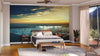 A modern bedroom features a wooden bed with gray bedding, matching nightstands, and a light area rug. Behind the bed is the Beach Panorama Wall Mural, depicting an ocean sunset. The room has hardwood floors, plants, and glass doors with sheer curtains.