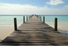 A partly cloudy sky arches over a wooden pier reaching into a turquoise ocean, flanked by weathered posts. This serene beach scene, with gentle waves kissing the shore, is reminiscent of the Beach Pier Wall Mural, an ideal peel and stick wallpaper design for tranquility.