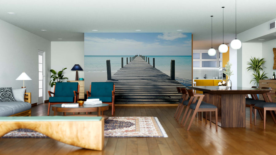 A modern open-plan living and dining room with wood furniture, blue chairs, a rug, and houseplants features the Beach Pier Wall Mural on the back wall, showing a wooden pier over turquoise water beneath a partly cloudy sky.