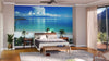 A spacious, modern bedroom features a bed centered against the Beach Scene Wall Mural. Wood floors, a ceiling fan, bedside tables with lamps, potted plants, and sheer curtains complete this sunlit retreat.