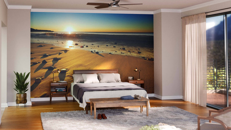 A cozy bedroom features a large Beach Sunset Wall Mural behind a wooden bed. Light walls, wood floors, a rug, bedside tables with lamps, a plant, and sheer curtains frame the glass door to the balcony.