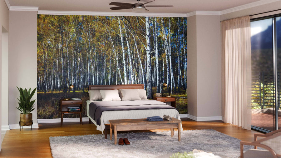 A modern bedroom boasts the Beautiful Autumn Sunny Day Wall Mural behind a wooden bed. Matching nightstands, bench, plants, and sheer curtains over glass doors complete the sunlit space with stylish ease.