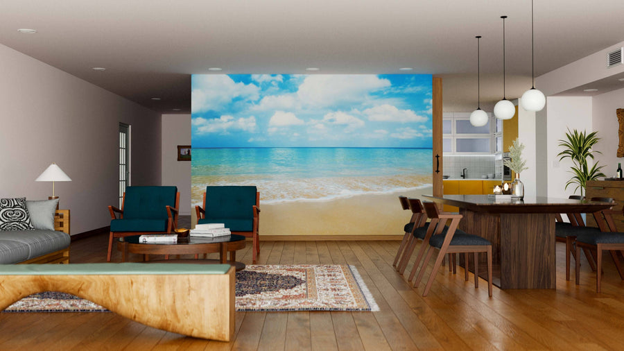 A modern open-plan living and dining area features wood floors, blue armchairs, a wooden table with chairs, and the Beautiful Beach Wall Mural for a bright beach vibe. Pendant lights hang above the table; a gray sofa and rug are in front.