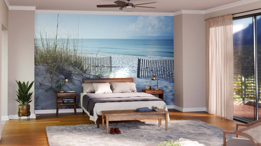 A bright bedroom features a bed with white bedding, gray blanket, wooden nightstands, lamps, a bench, and the Beautiful Beach in the Morning Wall Mural. Large windows with sheer curtains and a potted plant complete the airy space.