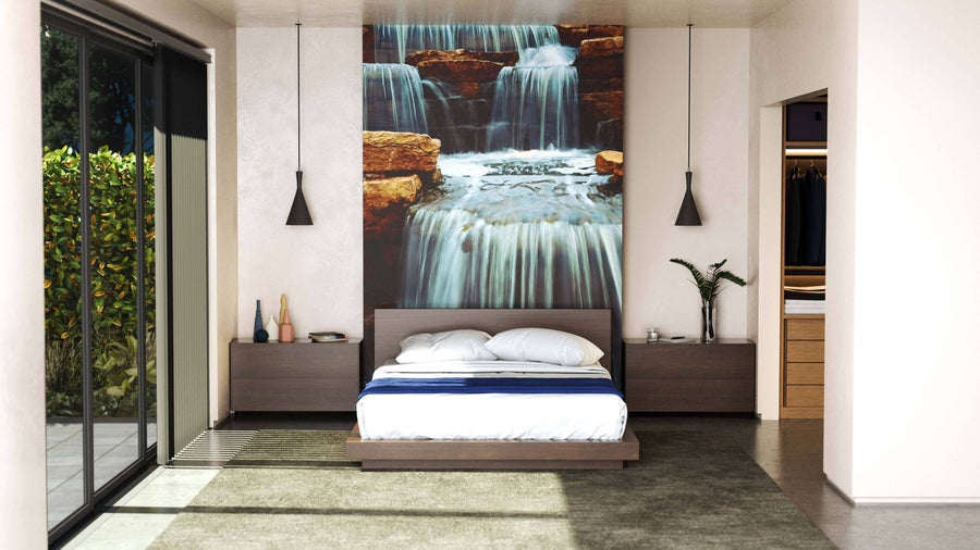 Modern bedroom featuring the Beautiful Cascading Waterfall Wall Mural behind a low wooden bed with white linens and a blue throw. Dark nightstands and pendant lights sit on either side; sunlight enters from a glass door, with a walk-in closet to the right.