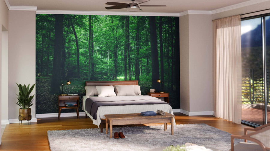 A modern bedroom features a Beautiful Green Forest Wall Mural behind the bed. The wooden bed with white bedding, matching nightstands, lamps, a bench, plant, rug, and sheer-curtained windows complete the serene space.