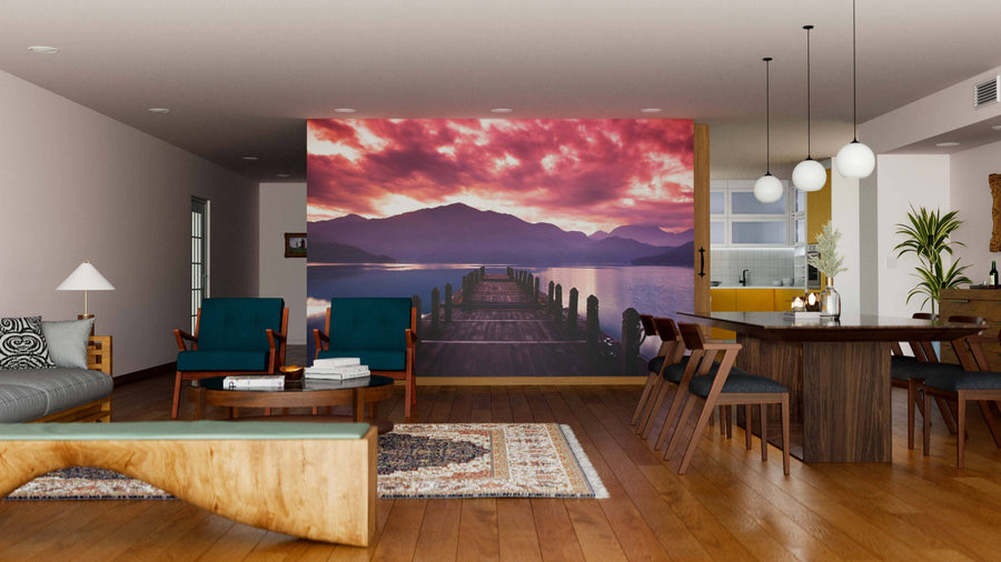 A modern living-dining room with wood floors, teal and wood furniture, a patterned rug, spherical pendant lights over the dining table, and a custom Beautiful Morning Sunrise Wall Mural depicting a pier over a lake at sunset with purple clouds.
