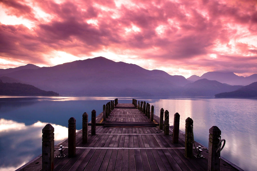 A modern living-dining room with wood floors, teal and wood furniture, a patterned rug, spherical pendant lights over the dining table, and a custom Beautiful Morning Sunrise Wall Mural depicting a pier over a lake at sunset with purple clouds.