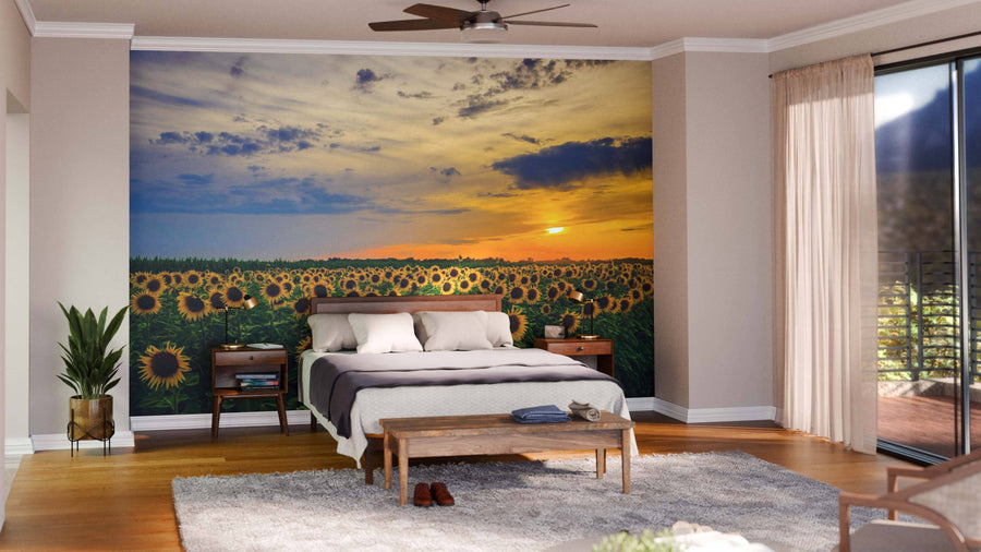 A bright bedroom showcases the Beautiful Sunflower Field Panorama Wall Mural behind a neatly made bed, with wooden floors, plants, a wooden bench, and large windows with sheer curtains welcoming natural light.