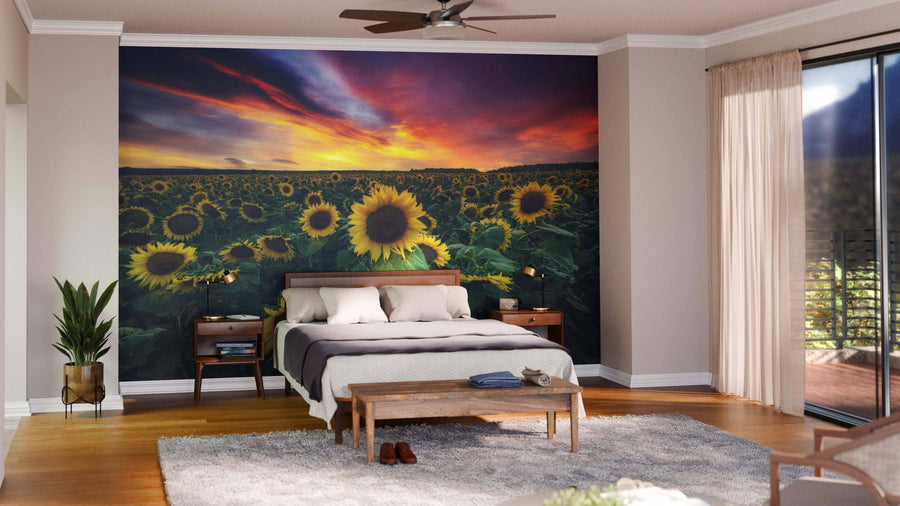 A cozy bedroom features a bed with white and gray bedding, a wooden bench, plants, and a rug. Behind the bed is the Beautiful Sunset over Sunflowers Field Wall Mural. Large windows fill the room with natural light.