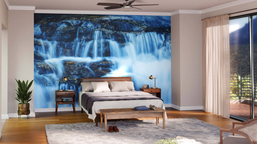 A modern bedroom features the Beautiful Waterfall Wall Mural behind the bed, wooden furniture, plants, a gray rug, and soft natural light streaming through sheer curtains and a glass door to the balcony. Light walls and a ceiling fan complete the space.