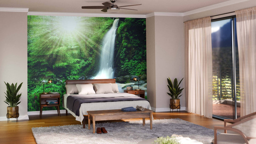 A cozy bedroom features the Beautiful Waterfall Czech Republic Wall Mural behind the bed. Wooden furniture, gray accents, soft rugs, and sliding glass doors create a warm, inviting space with sunlight streaming in.