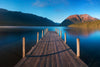 The serene "Between Peaks Wall Mural" features a long wooden dock over a calm, reflective lake surrounded by mountains under a clear sky with hints of clouds, perfectly capturing tranquility and natural beauty for your space.