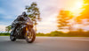 A motorcyclist in black gear speeds on a sleek, black sport motorcycle down a paved road. The blurred trees and vivid sunset evoke the motion of a Motor 18 Wall Mural, with partly cloudy skies in warm hues enhancing the dynamic scene.