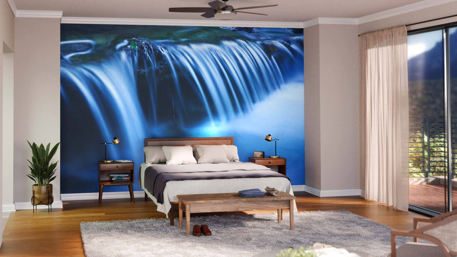 A modern bedroom features the Blue Cascade of Mountain River Wall Mural behind the bed, a wooden bed with white linens, two side tables with lamps, a bench at the foot, rug, potted plant, ceiling fan, and a sheer-curtained window.