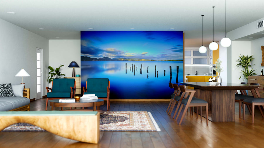 A modern open-plan living and dining area features the Blue Lake Wall Mural, depicting a serene lake with mountains and blue sky. Wooden furniture, green chairs, a gray sofa, pendant lights, and ample natural light enhance this inviting space.