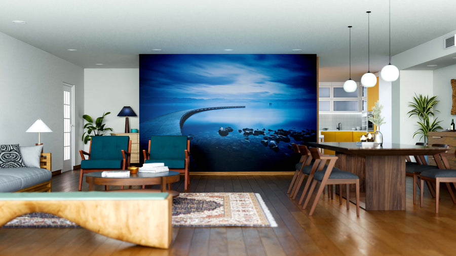 Modern open-concept living and dining room with wood floors, teal chairs, mid-century furniture, pendant lights, a patterned rug, green plants, a kitchen with yellow accents, and a Blue Sea and Curved Jetty Wall Mural as the focal point.