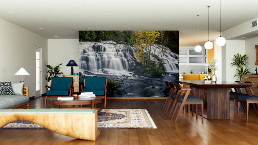 A modern open living-dining space features wood floors, blue chairs, a sofa, and a rug. The highlight is the stunning Bond Falls Wall Mural—a peel and stick mural of a cascading waterfall with lush greenery behind the dark wood dining table.