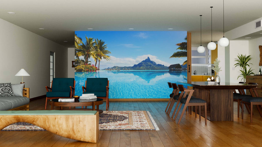 A modern living and dining room with wooden floors, mid-century furniture, and pendant lights features the Bora Bora Landscape Wall Mural, displaying a tropical pool with palm trees and mountains for an outdoor illusion.