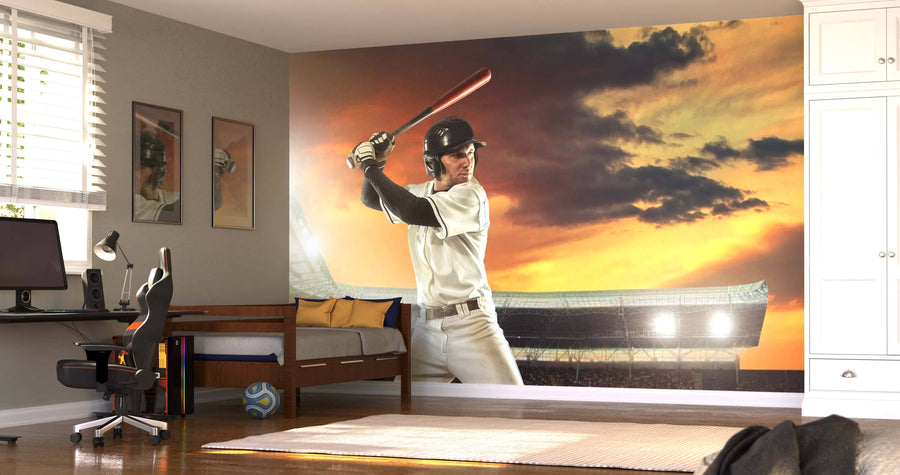 A modern bedroom showcases the Action Shot Wall Mural, depicting a baseball player swinging a bat at sunset. The room also has a bed, desk with computer, chair, window with blinds, photos on the wall, and a soccer ball on the wooden floor.