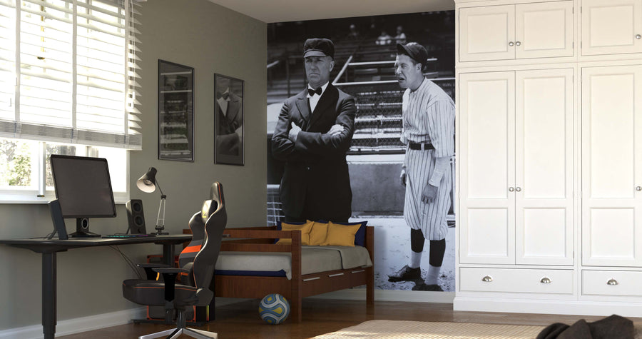 A modern bedroom with a desk, chair, and single bed features the Baseball Player and Umpire Wall Mural as a bold accent. Sunlight filters through white blinds; framed sports photos decorate the walls, and white built-in wardrobes line the right side.