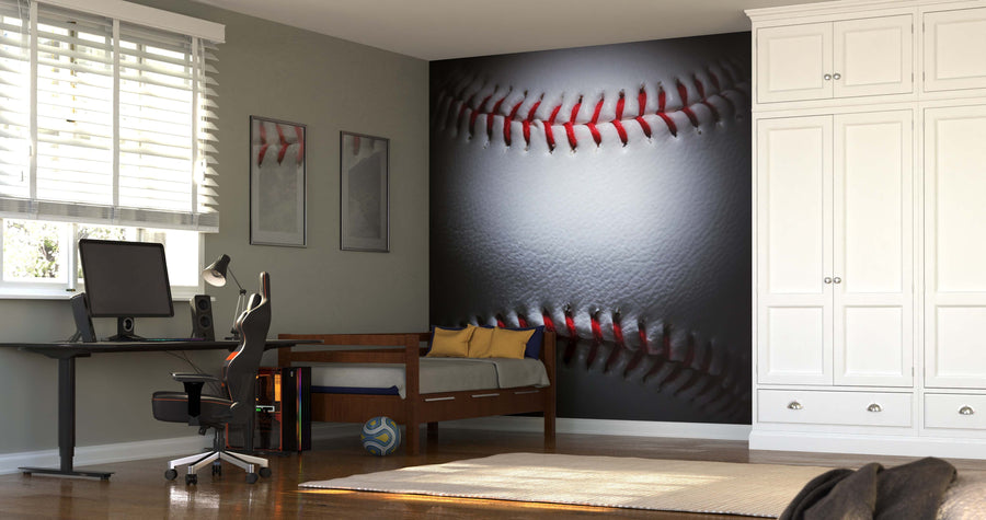 A bedroom features the Closeup of a Baseball Wall Mural, a modern desk with computer and chair, a daybed with yellow and gray pillows, wooden flooring, framed baseball art, a soccer ball, and white built-in wardrobes.