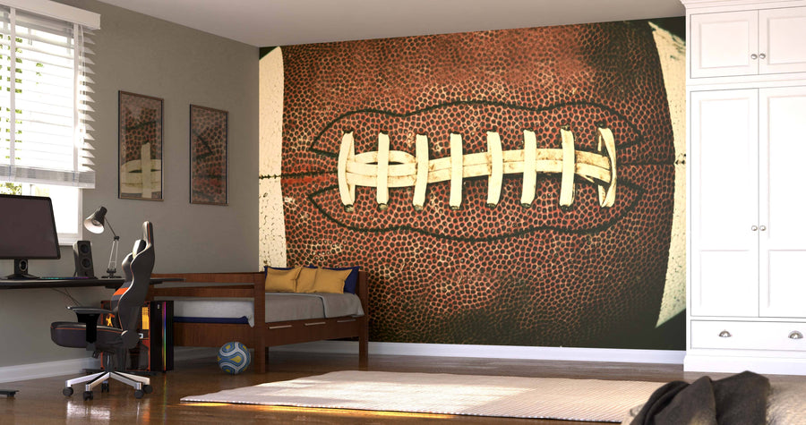 A modern bedroom showcases a large Football Wall Mural above a twin bed with yellow and gray pillows, alongside a desk, chair, wall art, wardrobe, and a football on the wooden floor. Sunlight filters through blinds.