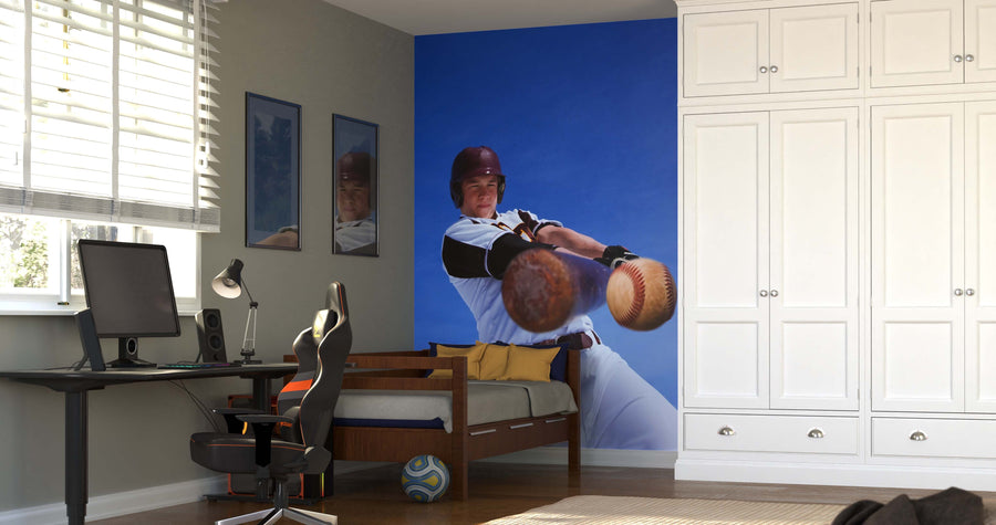 A modern bedroom features a desk, chair, bed, and the Home Run Hit Wall Mural—a large peel and stick design of a baseball player swinging a bat. White cabinets, framed pictures, and sunlight complete the fresh look.