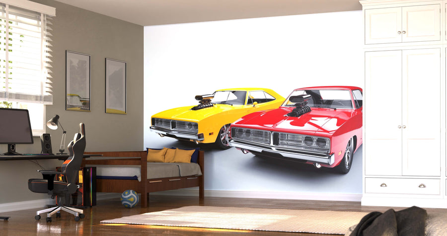 A modern bedroom showcases the Muscle Cars Wall Mural, displaying two vintage muscle cars—one yellow and one red—side by side. The room also features a bed, desk with computer, black chair, ball on the floor, white cabinets, and sunlight.