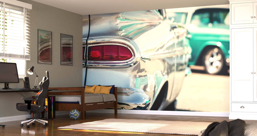 A modern bedroom features a desk, chair, and bed with yellow pillows. Behind the bed, the Old American Car Wall Mural with a shiny chrome tail fin and red tail light creates a vintage vibe. Sunlight streams through the blinds.