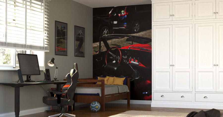 A modern bedroom with a white wardrobe, computer desk, black gaming chair, and a bed with blue and yellow pillows. The Red Vintage Car Wall Mural adds flair as sunlight streams in, highlighting the wooden floor and soccer ball.