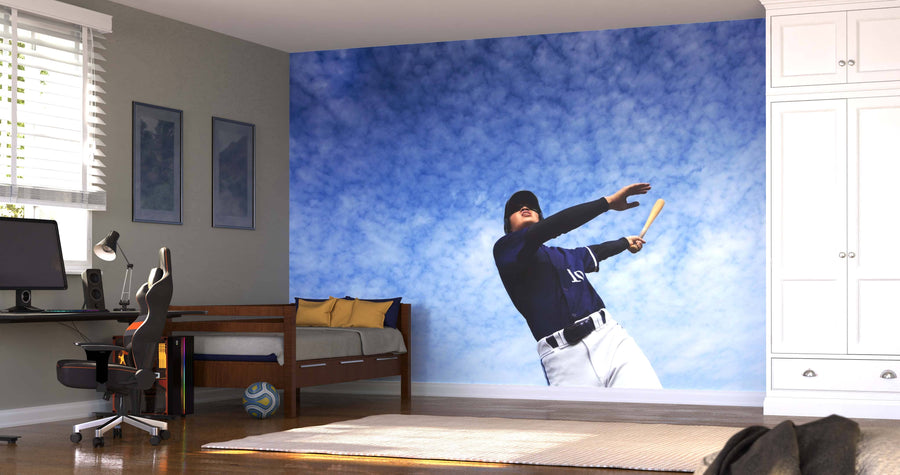 A tidy bedroom features the Swing for the Sky Wall Mural of a baseball player swinging a bat, a desk with computer and chair, bed with yellow pillows, soccer ball, two paintings, wooden floor, and white wardrobe.